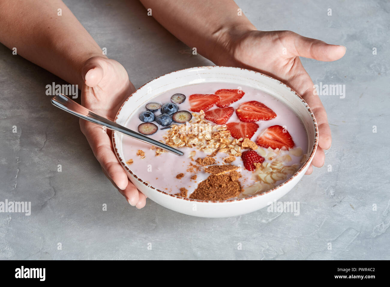 Frau Hände halten Schüssel mit appetitlich gesund Müsli Nachtisch mit Stücken von Erdbeeren, Sahne, Haferflocken auf grauem Beton Hintergrund Stockfoto