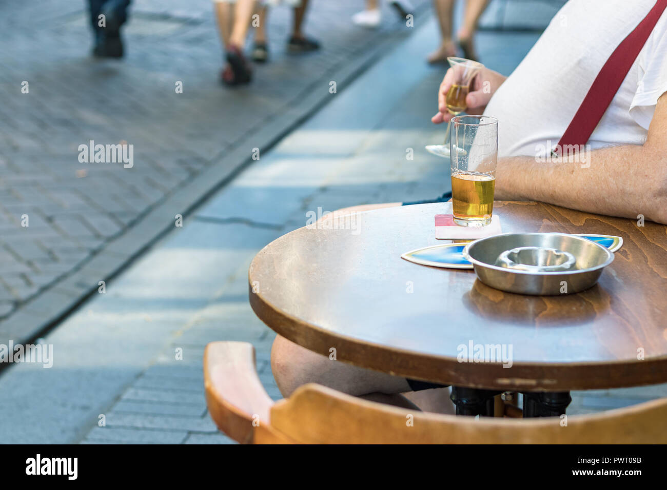 Ein Mann mit großen Bauch sittiing draußen in einer Bar mit einem Arm auf dem runden Holztisch Bier trinkt. Stockfoto