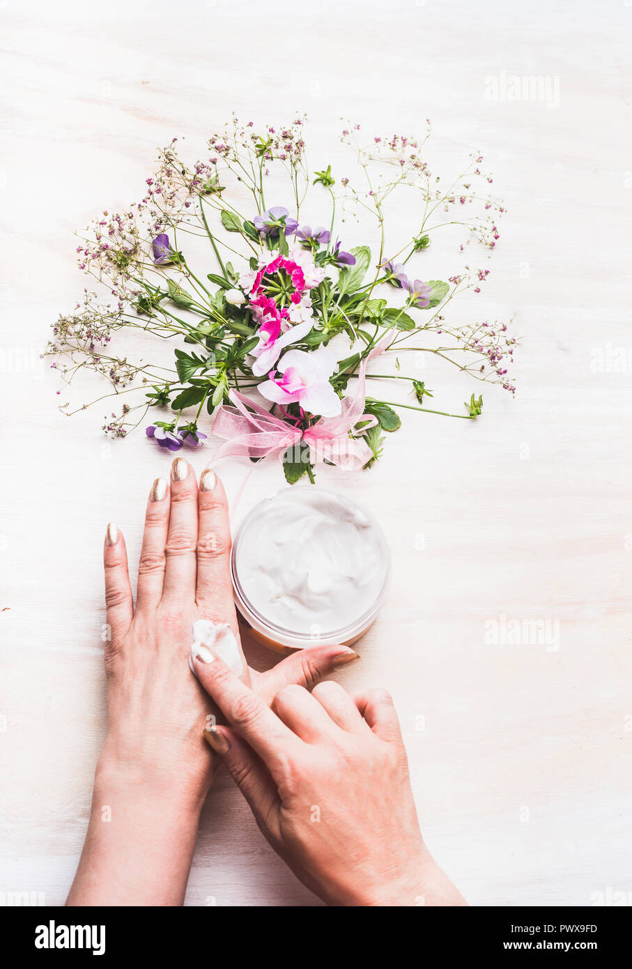 Weibliche Hände tragen Sie die Creme auf die Haut auf weißem Hintergrund mit Pflanzen und Blumen, Ansicht von oben. Natürliche Kosmetik Konzept Stockfoto