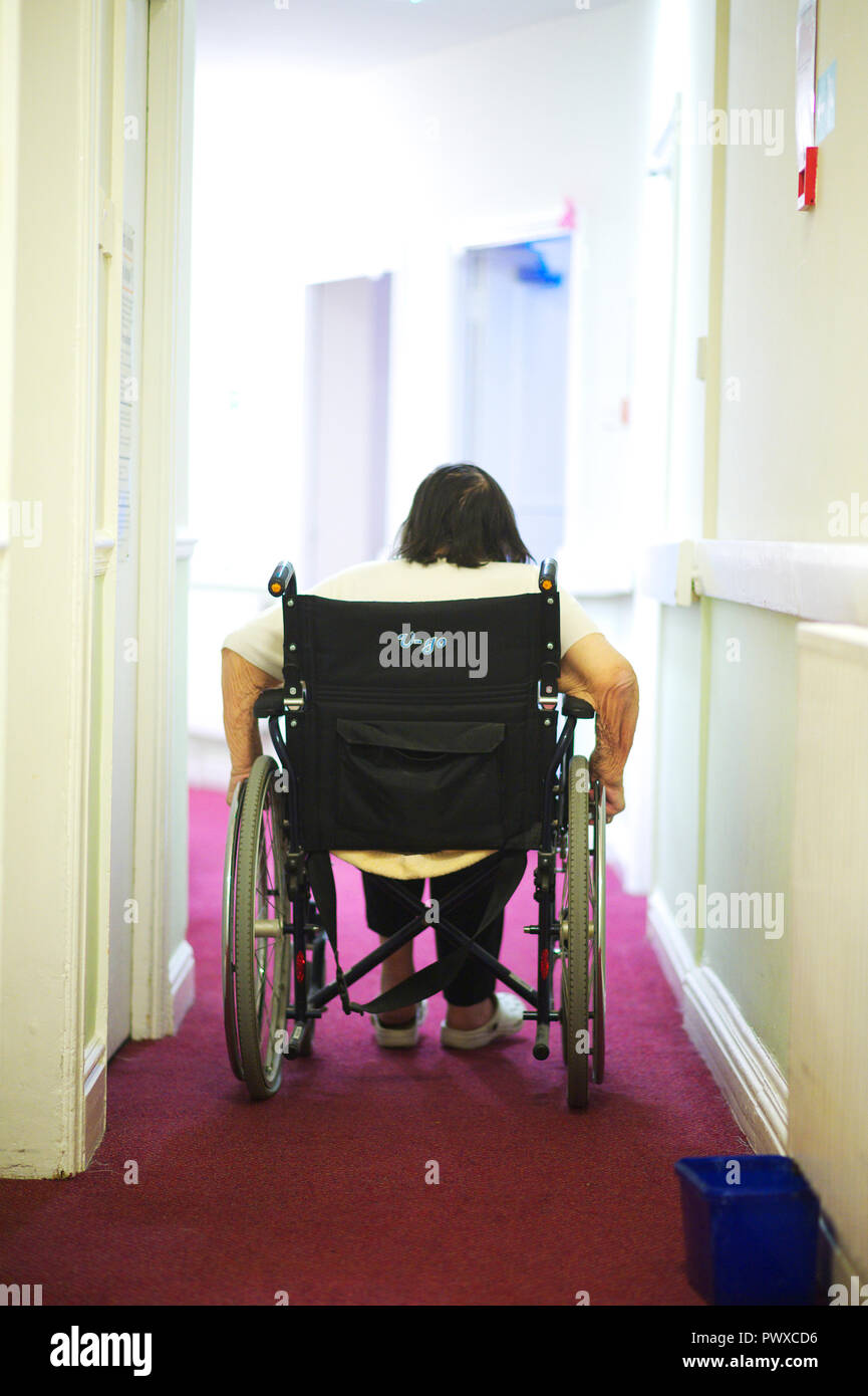 Die älteren behinderten Frau selbst in einem Rollstuhl mit einem Korridor in einer Regierung care Home drücken Stockfoto