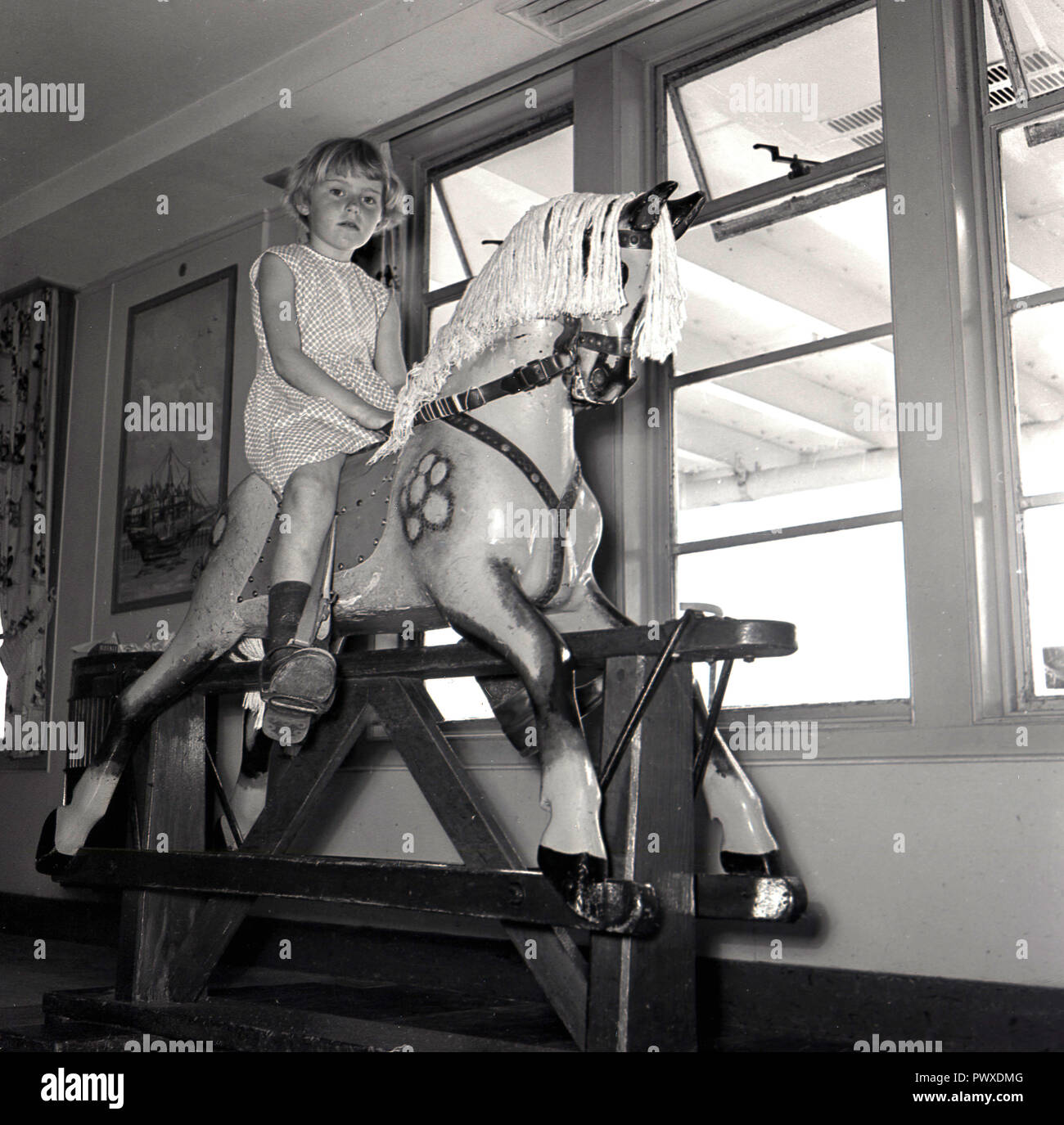 1950, historische, auf einem Dampfschiff, in den Kindergarten oder Spielplatz, ein junges Mädchen sitzt mit ihren Füßen in der stirups spielen oder Reiten auf einem hölzernen Schaukelpferd am Fenster. Stockfoto