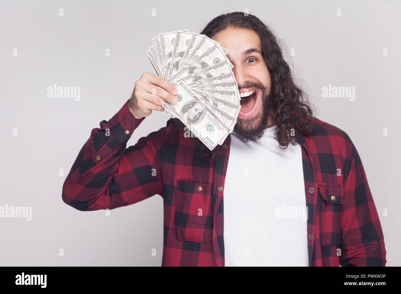 Junge Unternehmer in rot kariertem Hemd und lange lockige dunkle Haare stehen erstaunt, hält das Gebläse von Dollar und für Auge und mit überrascht fac Gesicht Stockfoto