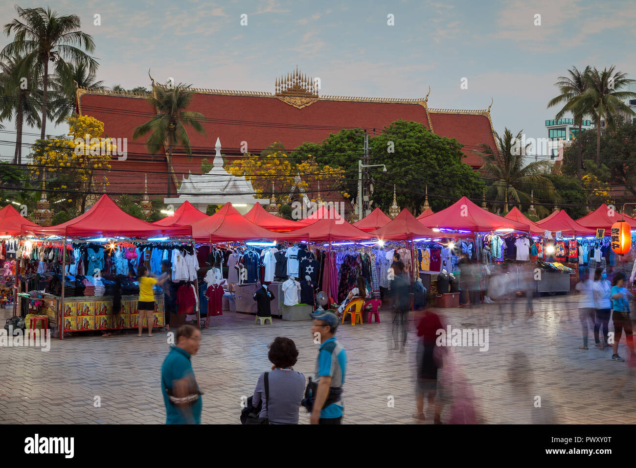 Menschen und viele Stände voller Kleider an der berühmten Nachtmarkt in Vientiane, Laos, in der Dämmerung. Wat Chanthaburi (Chanthaboury Tempel im Hintergrund ist. Stockfoto