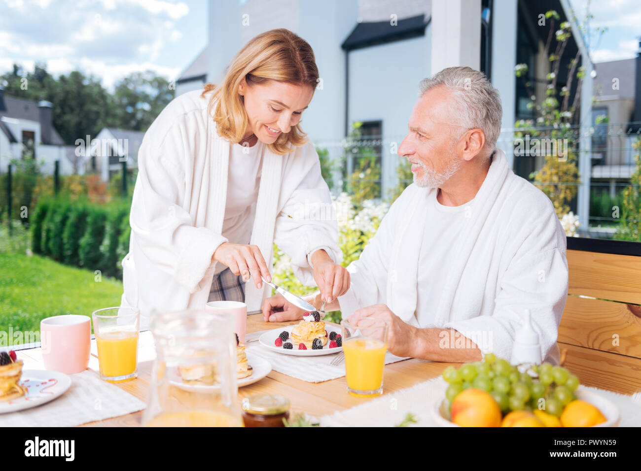 Liebevolle gut aussehende Frau schneiden Morgen Pfannkuchen mit Früchten für ihren Mann Stockfoto