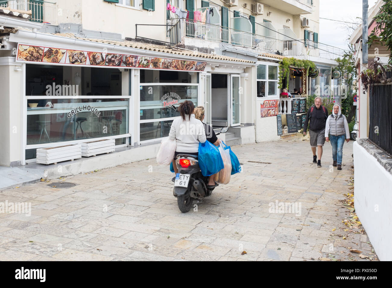 Zwei Frauen reiten Sozius auf einem Motorrad ohne Helm und Durchführung Shopping auf einer schmalen Straße in Kassiopi, Korfu Stockfoto