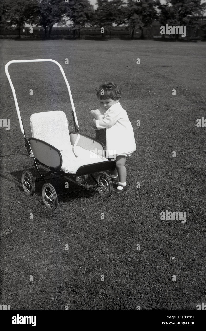 1950, historische, kleine Mädchen im Freien in einem Park mit ihren weichen Spielzeug, neben Kinderwagen ihres Metall Puppe oder Spielzeug Kinderwagen, England, UK. Stockfoto