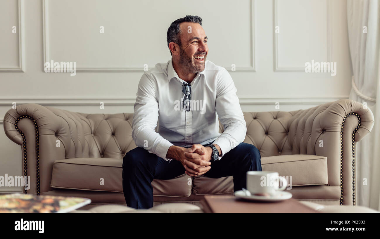 Portrait von fröhlichen Geschäftsmann sitzt auf einem Sofa im Hotel Zimmer und Wegsehen lachen. CEO der Aufenthalt in einem luxuriösen Hotel auf einer Geschäftsreise. Stockfoto