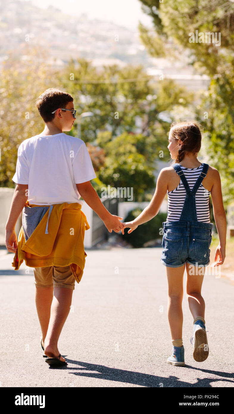 Ansicht der Rückseite ein Junge und ein Mädchen zu Fuß auf der Straße zu suchen. Kinder gehen zusammen auf der Straße ihre Hände halten an einem sonnigen Tag. Stockfoto