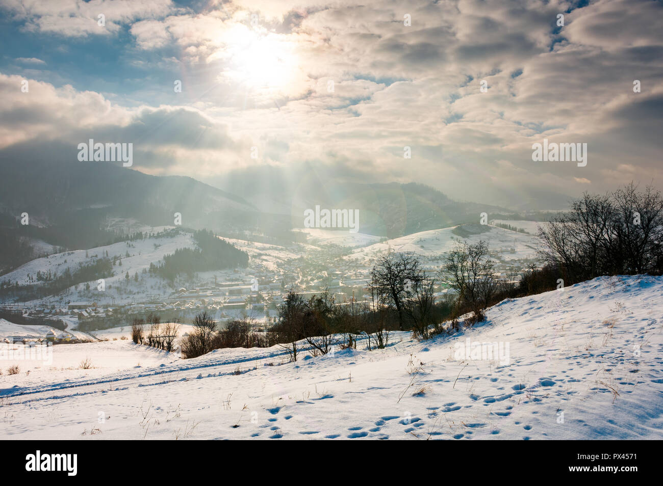 Zauberhafte Winterlandschaft. Sun Ray durch den bewölkten Himmel. verschneite Hügel und kahlen Bäumen. Dorf unten im Tal Stockfoto