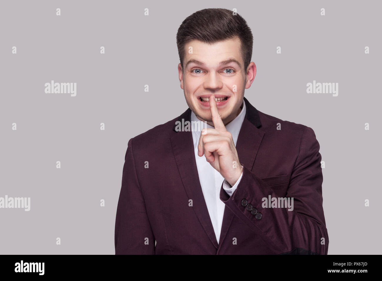Shh, ihren Willen zwischen uns. Lustige smiley hübscher junger Mann in violett Anzug, weißes Hemd, stehend, mit Blick auf die Kamera mit den leisen Zeichen und sagt sec Stockfoto