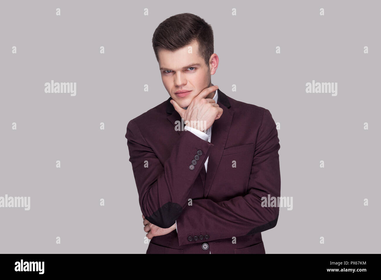 Portrait von Stattlichen ernsthaften jungen Mann in violett Anzug und weißes Hemd, stehend, sein Kinn berühren und Kamera mit nachdenklichen Gesicht. indoor Stockfoto