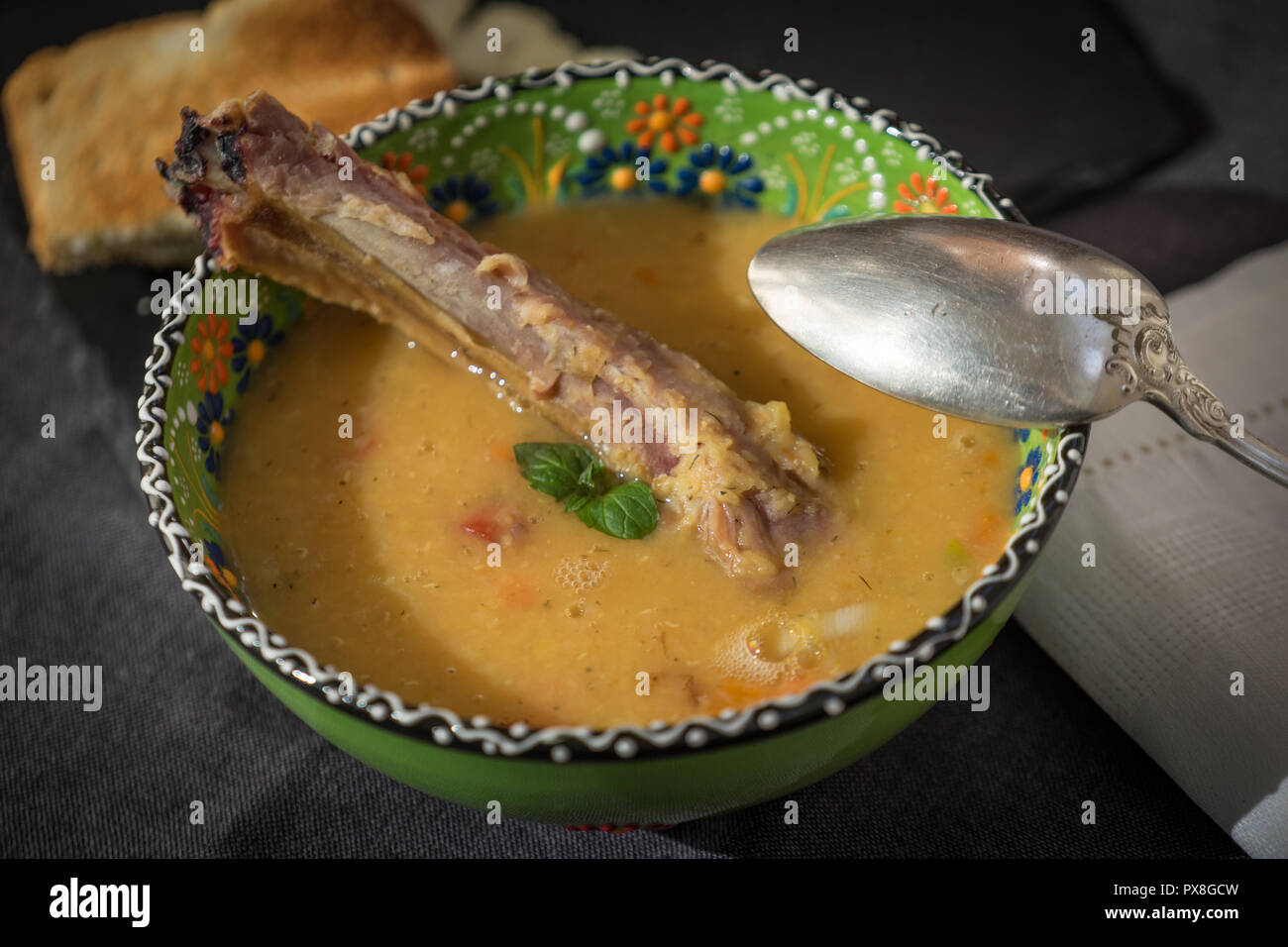 Rote Linsensuppe mit geräuchertem Rippchen in einem hellen Grün Schüssel mit floralen Ornament. Dunkelgrau Stoff Hintergrund. Stockfoto