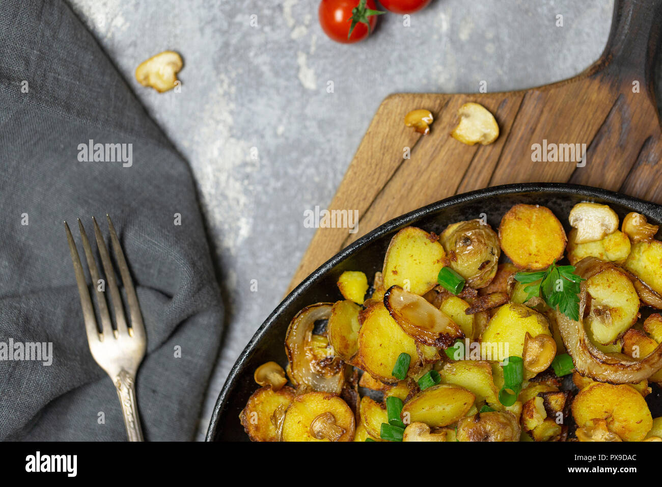 Rustikale frische Bratkartoffeln mit Pilzen und Zwiebeln in eiserne Pfanne mit Gabel, frische Tomaten, gebratene Champignons auf Holz Board am grauen backgroun Stockfoto