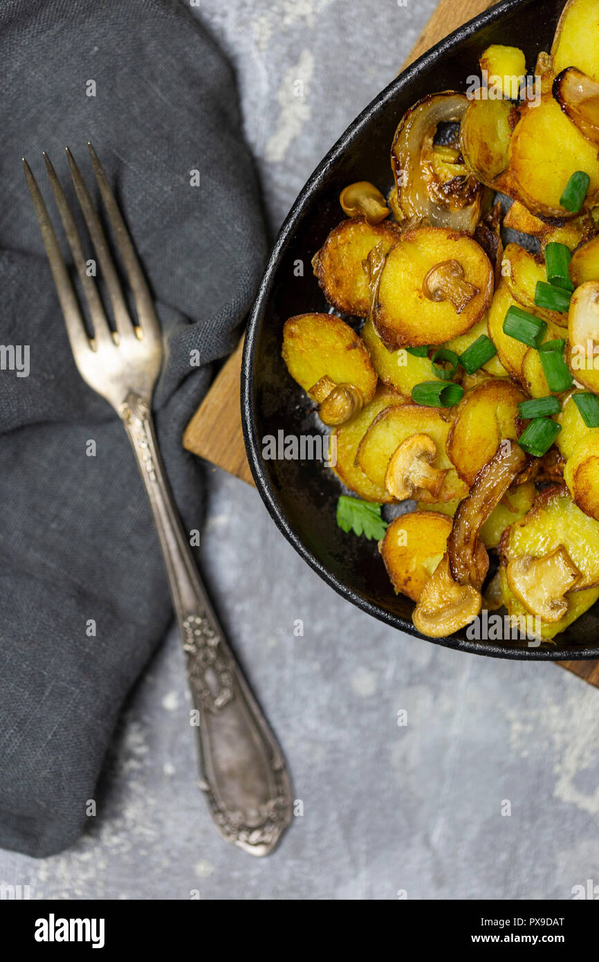 Rustikale frische Bratkartoffeln mit Pilzen und Zwiebeln in eiserne Pfanne mit Gabel, frische Tomaten, gebratene Champignons auf Holz Board am grauen backgroun Stockfoto
