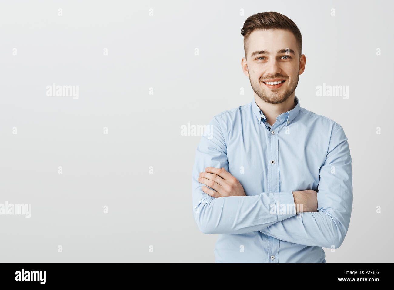 Selbstbewusst und freundlich aussehenden Mann mit Borsten in Blue Collar Shirt, Hände auf der Brust gekreuzt in self-assured pose Lächeln freudig intirigued neue Idee zu hören, wie Sie Kunden gewinnen Stockfoto
