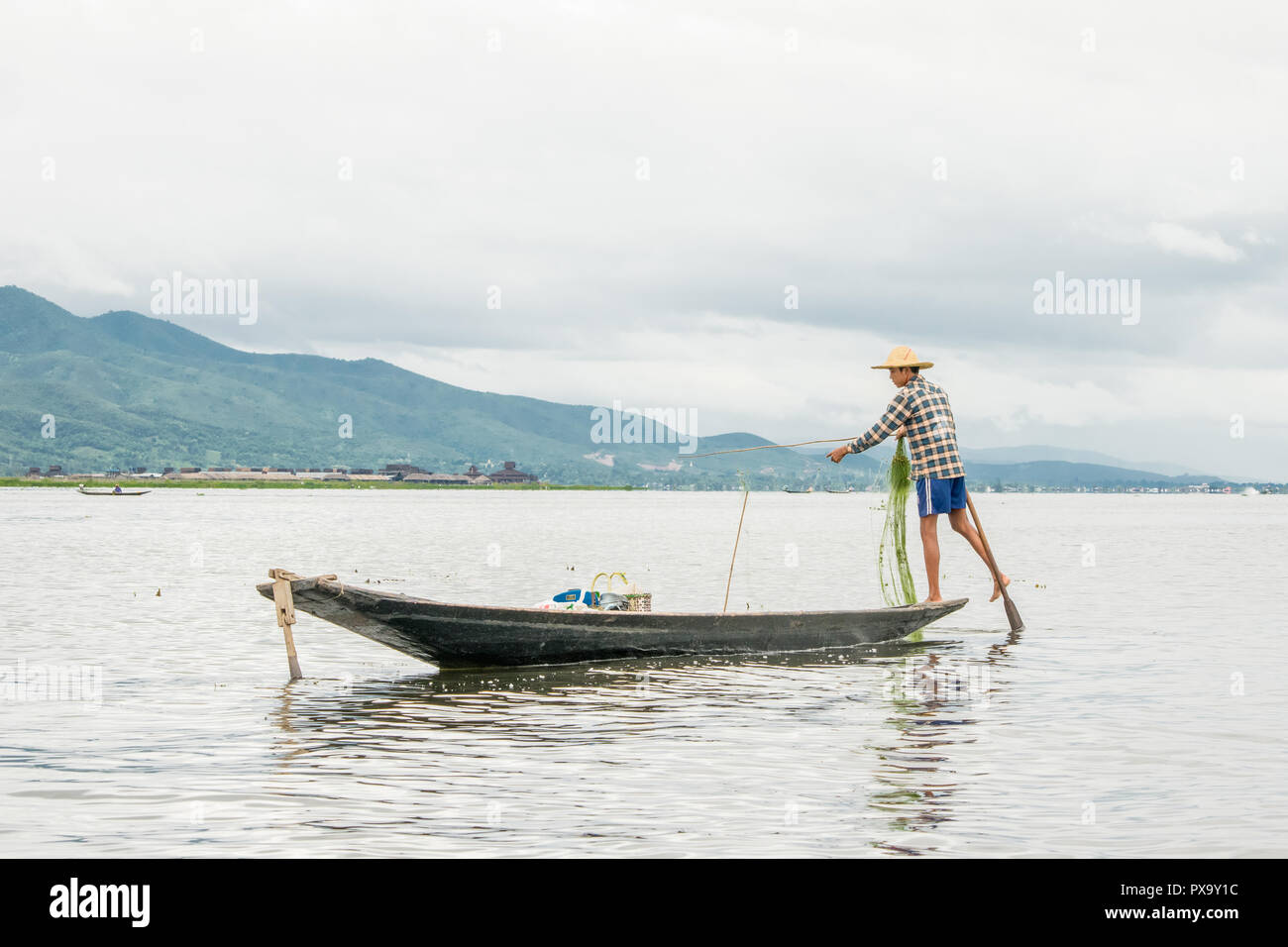 Reisen lokale junge burmesen männliche Fischer tragen geprüft, t-shirt, mit Stick und net zu fischen, Balancieren auf einem Fuß auf dem Boot, Inle Lake Myanmar, Birma Stockfoto