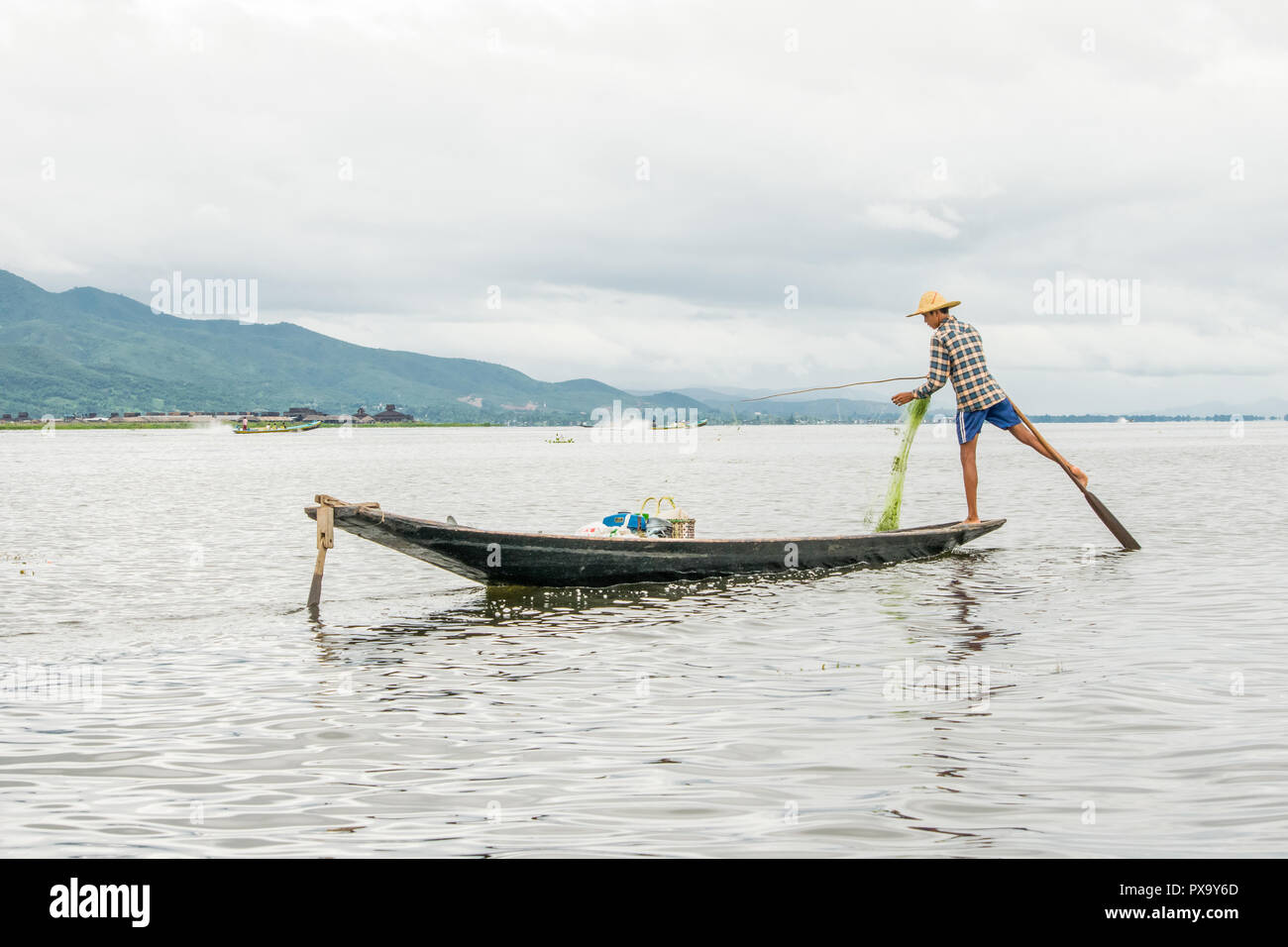 Reisen lokale junge burmesen männliche Fischer tragen geprüft, t-shirt, mit Stick und net zu fischen, Balancieren auf einem Fuß auf dem Boot, Inle Lake Myanmar, Birma Stockfoto