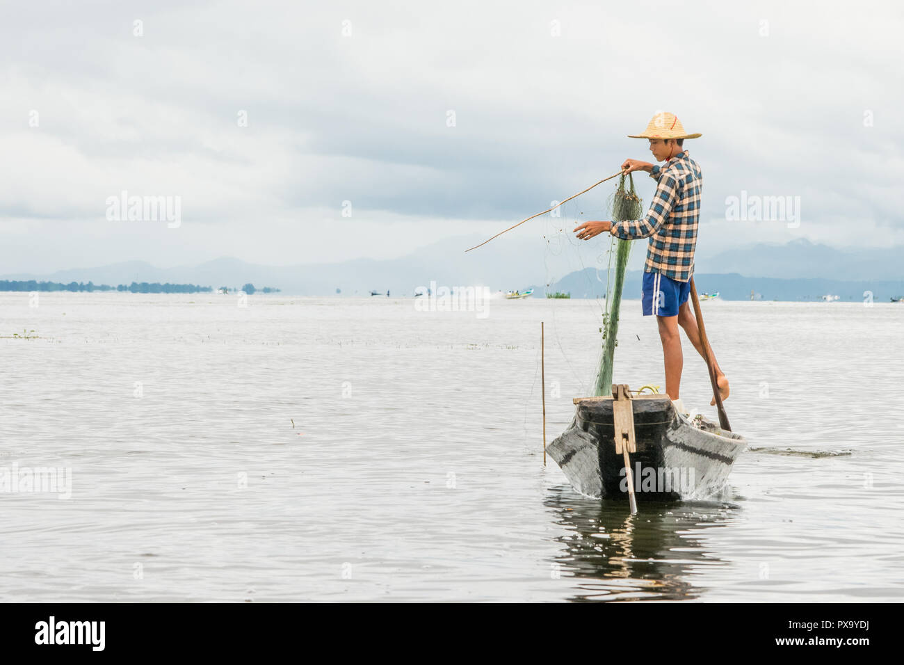 Reisen lokale junge burmesen männliche Fischer tragen geprüft, t-shirt, mit Stick und net zu fischen, Balancieren auf einem Fuß auf dem Boot, Inle Lake Myanmar, Birma Stockfoto