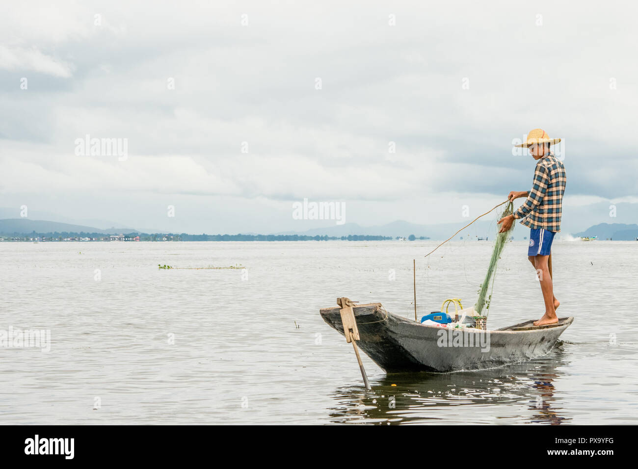 Reisen lokale junge burmesen männliche Fischer tragen geprüft, t-shirt, mit Stick und net zu fischen, Balancieren auf einem Fuß auf dem Boot, Inle Lake Myanmar, Birma Stockfoto