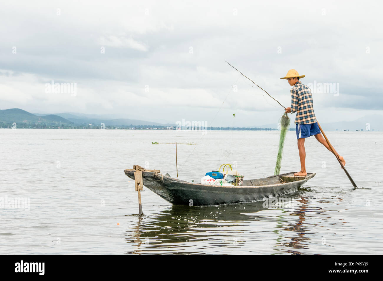 Reisen lokale junge burmesen männliche Fischer tragen geprüft, t-shirt, mit Stick und net zu fischen, Balancieren auf einem Fuß auf dem Boot, Inle Lake Myanmar, Birma Stockfoto