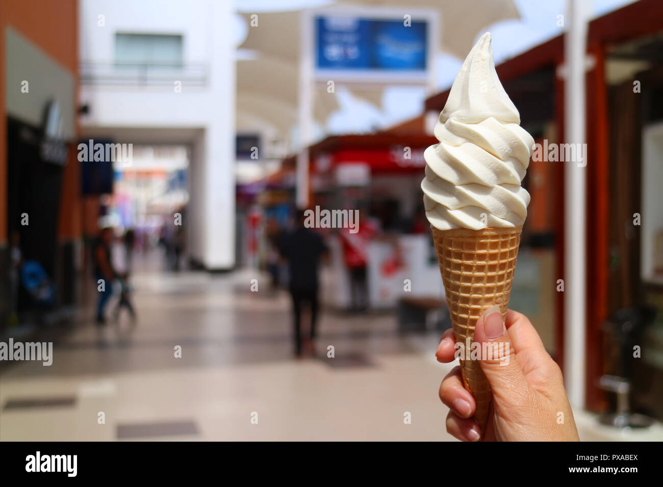 Woman's Hand, die Vanille Milch weiche Eis in der Sonne dienen, mit unscharfen Shopping Mall im Hintergrund Stockfoto