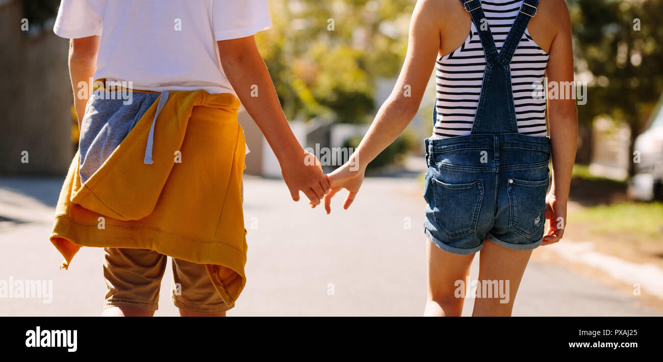 7/8-schuss ein Junge und ein Mädchen zu Fuß auf der Straße halten sich an den Händen an einem sonnigen Tag. Kinder in der Liebe zu Fuß auf eine leere Straße halten sich an den Händen. Stockfoto