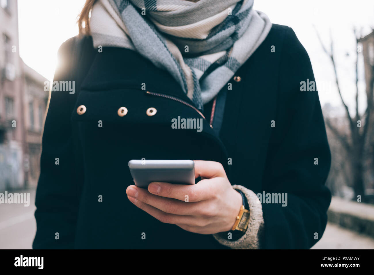 Junge Frau mit Smart Phone im Freien auf grauen Hintergrund. Frauen tragen große Schal, gold Armbanduhren, schwarzes Fell und Rucksack Holding mobile d Stockfoto