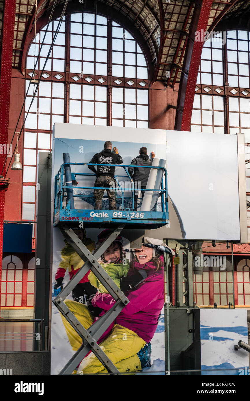 Antwerpen, Belgien - 2018-10-01: Hoch über einem belebten Bahnhofshalle, auf einem highworker Gerüst Plattform auf Rädern, zwei Männer beschäftigt sind, einen riesigen advertisem Stockfoto