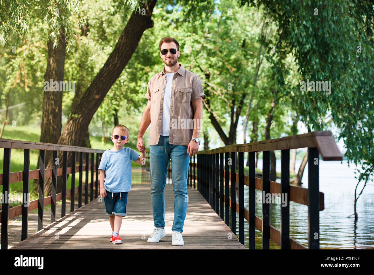 Vater und Sohn in Sonnenbrille halten sich an den Händen und laufen auf der Brücke im Park Stockfoto