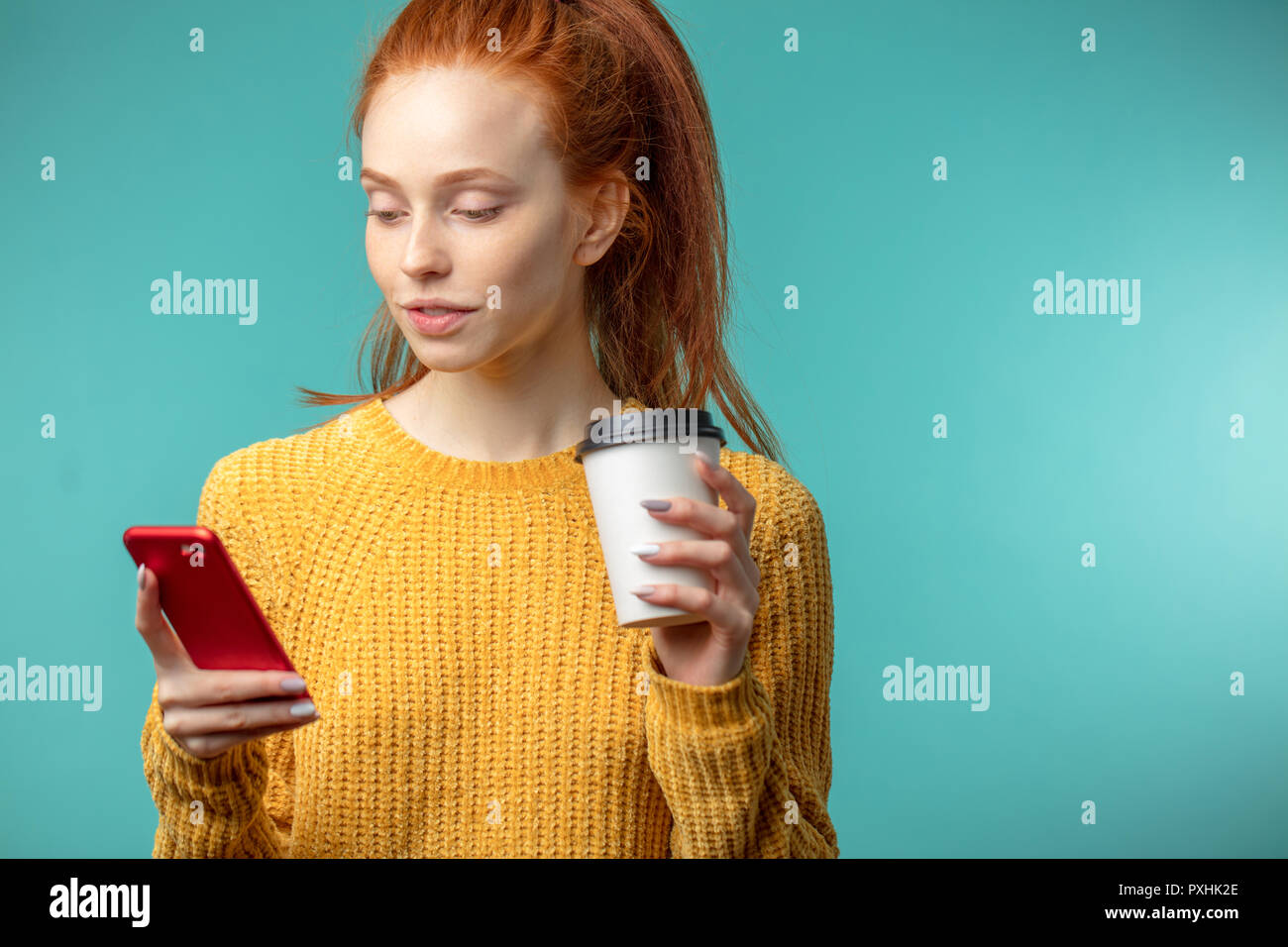Junge rothaarige Frau mit Kaffee und Smartphone über blauen Hintergrund isoliert Stockfoto