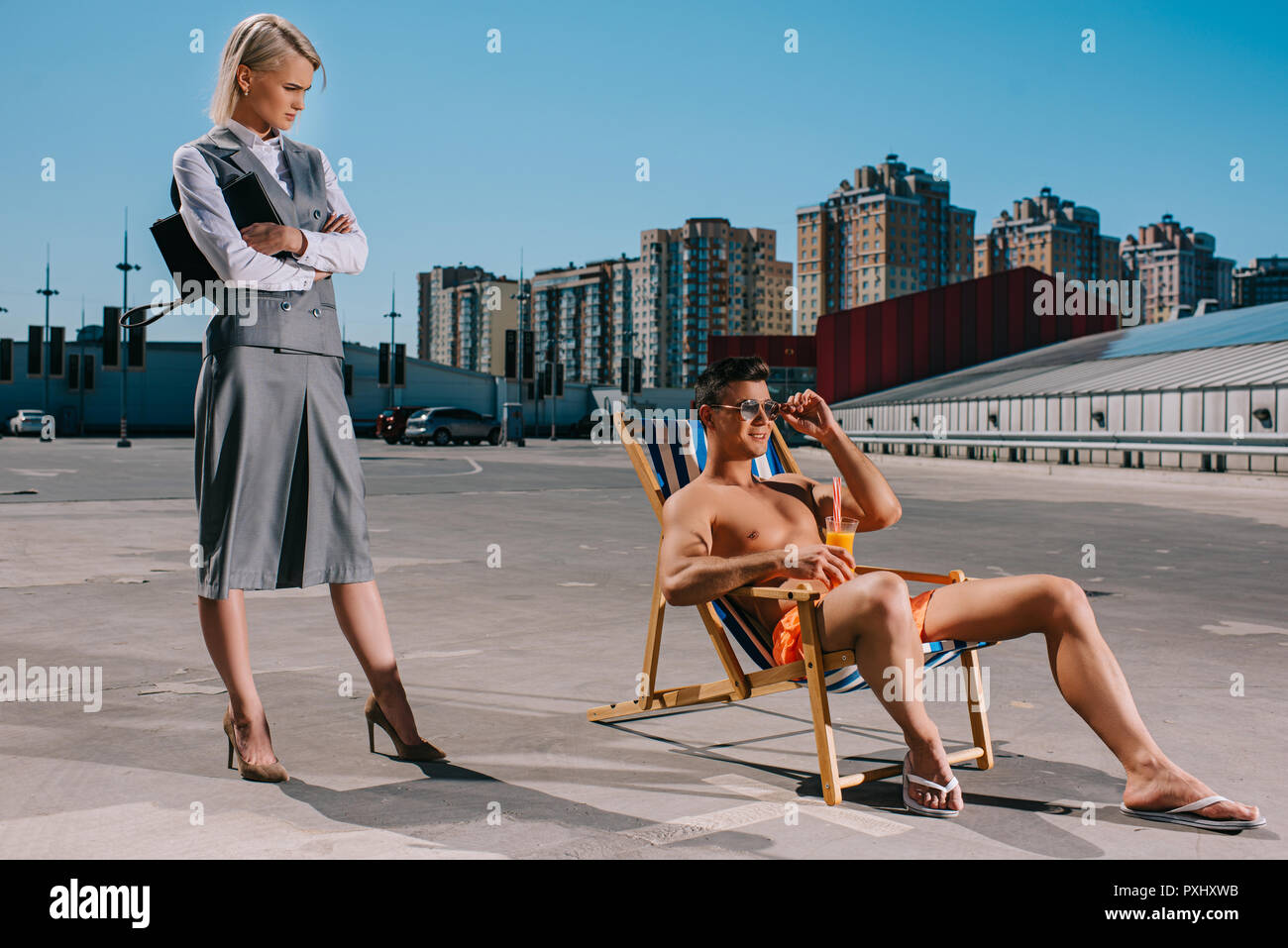 Hübscher junger Mann relaxen im Liegestuhl, während seine Frau Chef im Anzug neben ihm mit verschränkten Armen auf den Parkplatz Stockfoto