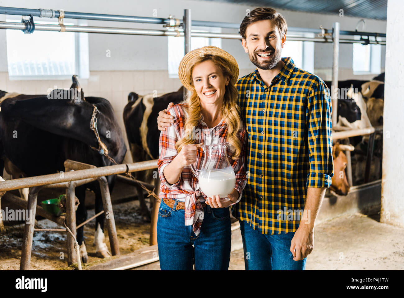 Lächelndes Paar Landwirte halten Kanne Milch in Stall mit Kühen Stockfoto