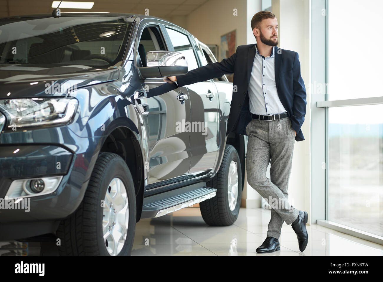 Stattliche ernster Mann stehen und in der Nähe von dunklen blauen Jeep und große Fenster im Auto Center posieren. Unternehmer tragen in dunkel blau Jacke und blaue T-Shirt. Kunden Einkauf neue Auto. Stockfoto