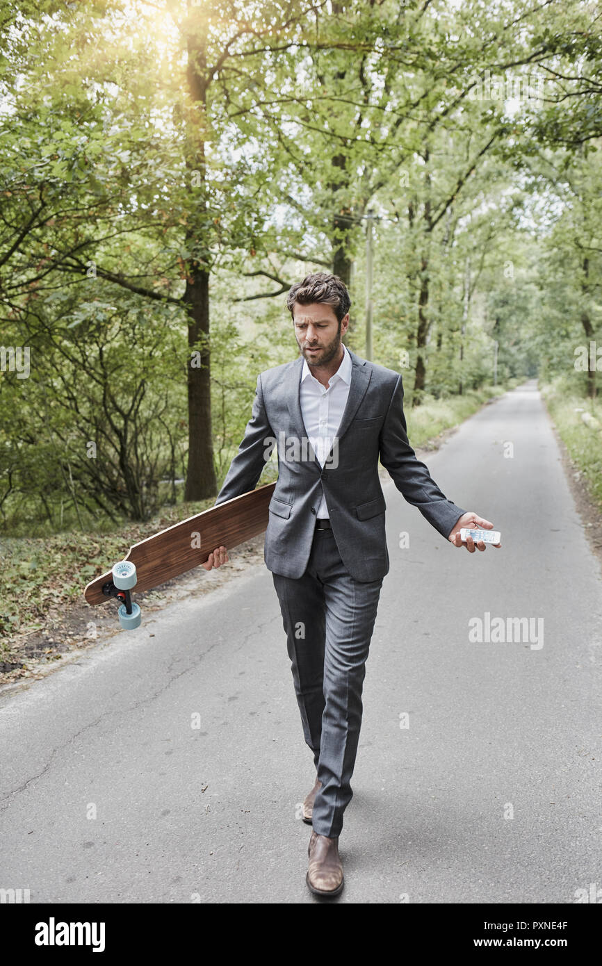 Unternehmer gehen mit Skateboard und Smartphone auf die ländliche Straße Stockfoto