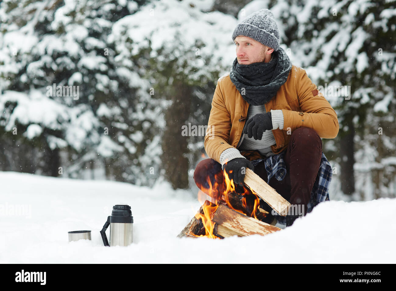 Nachdenklich hübscher junger männlicher Wanderer in warmer Schal am Lagerfeuer zu sitzen, während es die Beleuchtung und das Hinzufügen von Hölzer im Winter Wald, er schaut in die Ferne Stockfoto
