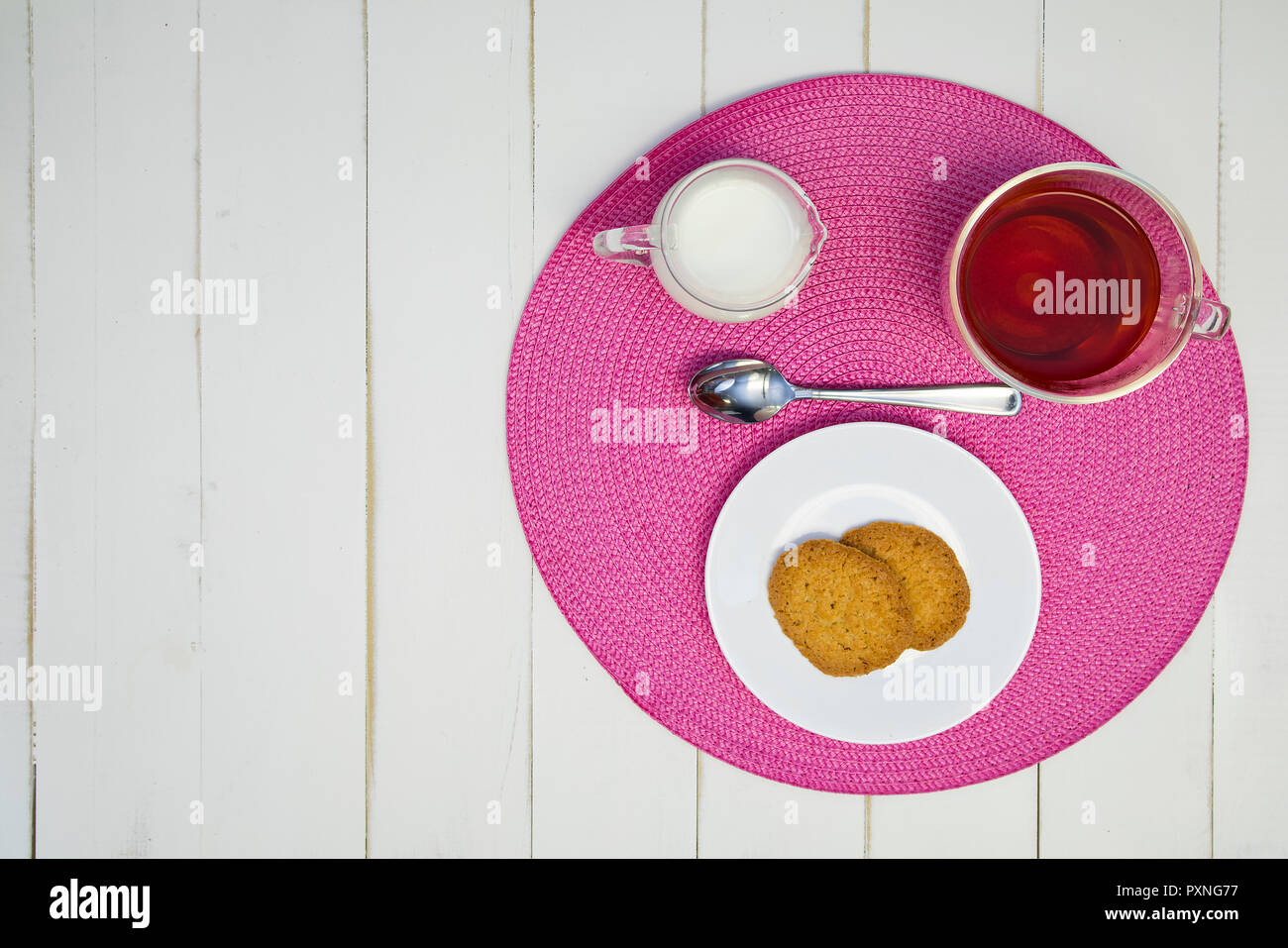 Heißen Tee, Kekse und Milch sind auf einem rosa Tischset auf eine weiße Tafel Tabelle angeordnet. Die Anordnung ist off-center. Stockfoto