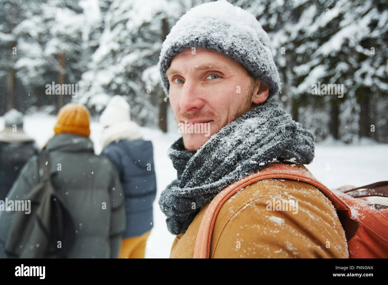 Inhalt hübscher junger männlicher Wanderer in verschneiten Hut und Schal zurück zu drehen, während an der Kamera im Winter Wald suchen, er folgende Gruppe von Reisenden Stockfoto
