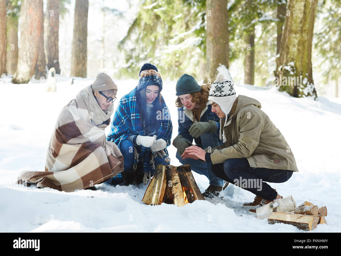 In voller Länge Porträt von vier Jugendlichen Camping im Winter Wald im Kreis um das Feuer sitzen Stockfoto