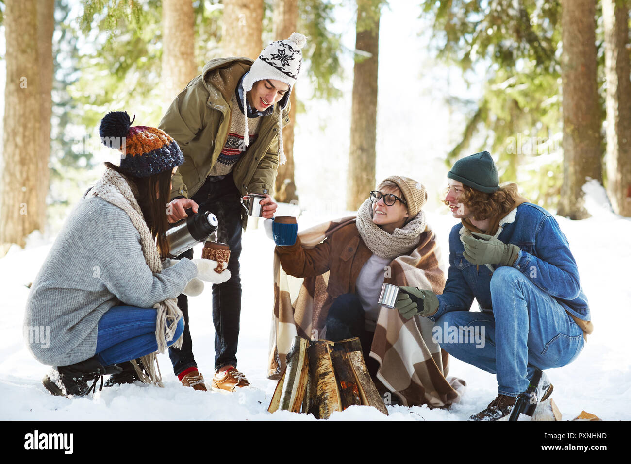 In voller Länge Porträt von vier Jugendlichen Camping im Winter Wald im Kreis um das Feuer sitzen und strömenden heißen Getränken Stockfoto