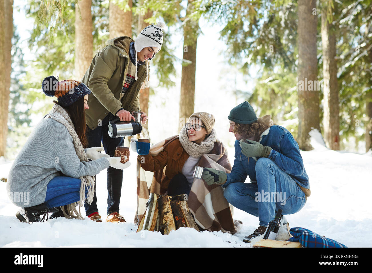 In voller Länge Porträt von vier jungen Freunde Camping im Winter Wald im Kreis um das Feuer sitzen und strömenden heißen Getränken Stockfoto