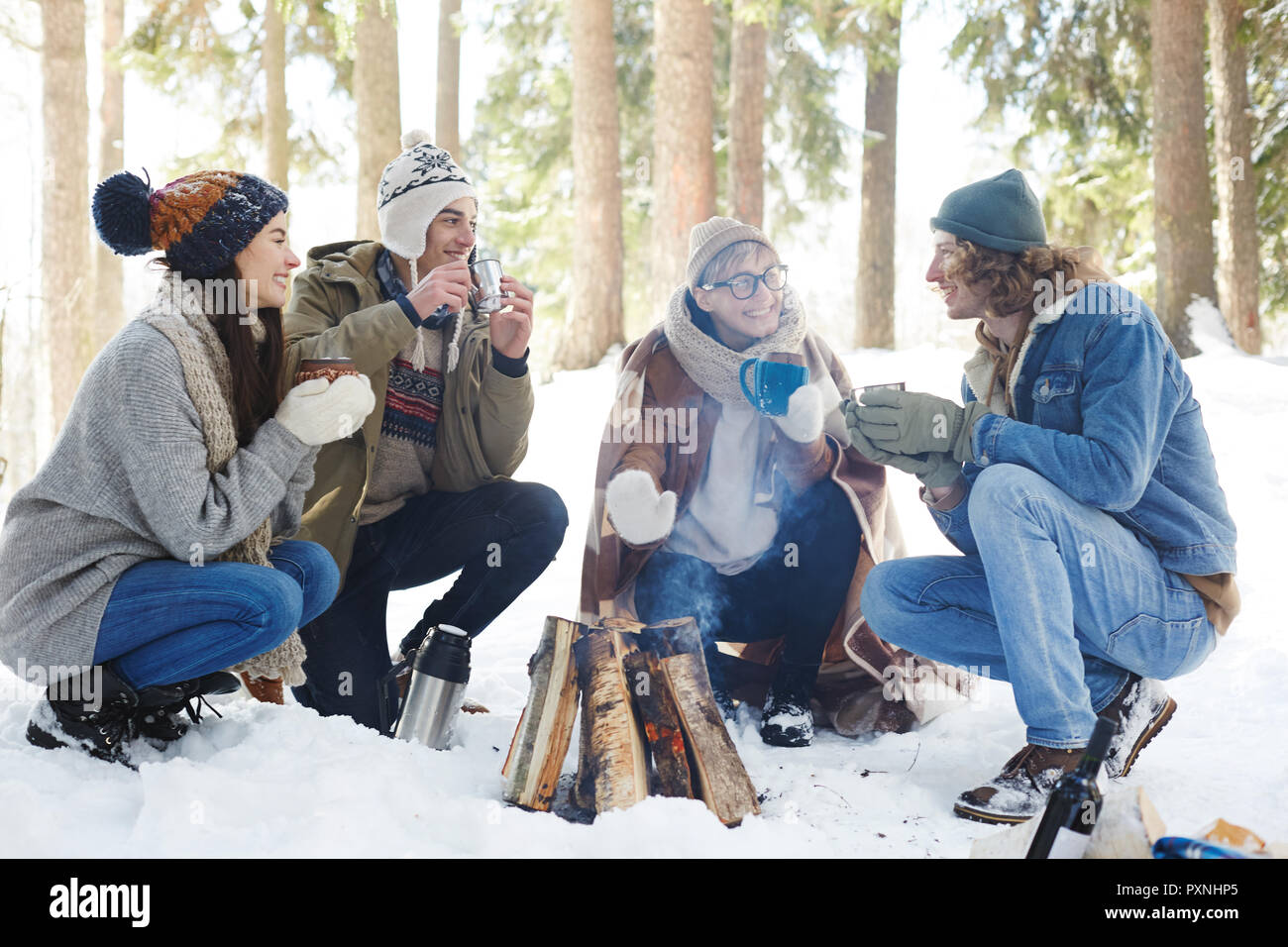 In voller Länge Porträt von vier Jugendlichen Camping im Winter Wald im Kreis um das Feuer sitzen und plaudern beim Trinken von heißem Kakao Stockfoto
