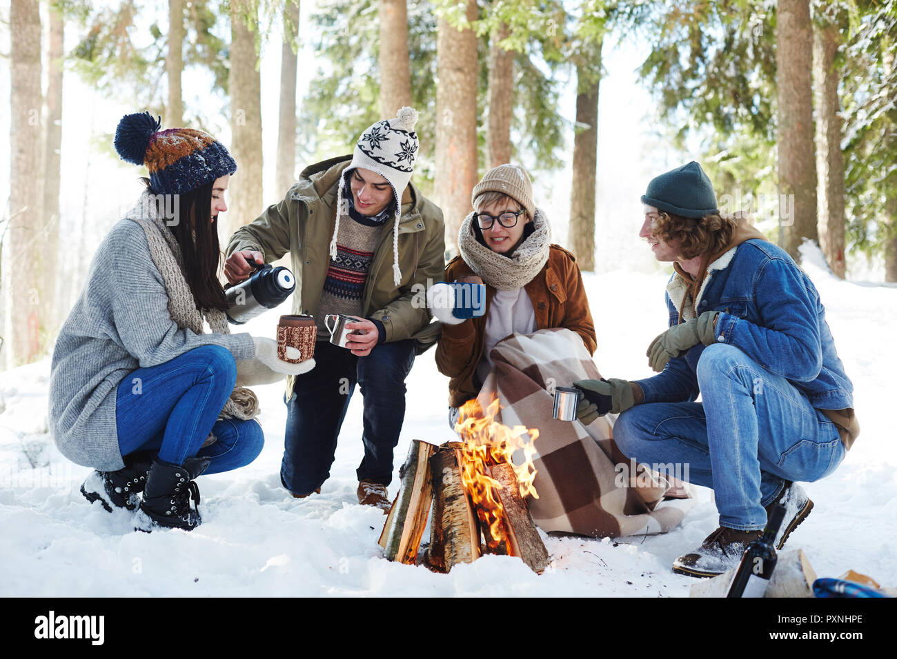 In voller Länge Porträt von vier Jugendlichen Camping im Winter Wald im Kreis um das Feuer sitzen und trinken heißen Kakao Stockfoto