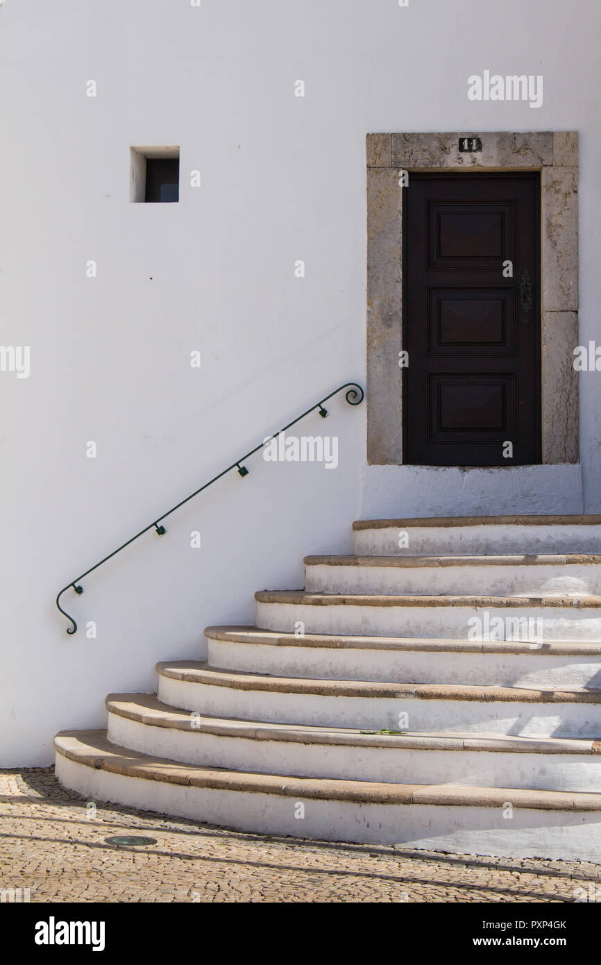 Gepflasterte Gasse mit Kopfsteinpflaster. Gebogene Treppe und eine Tür in einem Rahmen. Weißen hellen Fassade des Gebäudes. Stilvolle Griff an der Wand. Faro, Portugal. Stockfoto