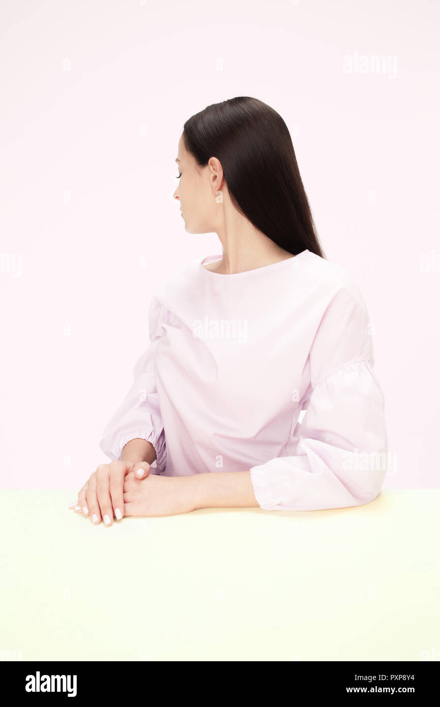 Serious Business Frau wandte sich am Tisch sitzen auf einem Pink Studio Hintergrund. Das Porträt in minimalistischem Stil Stockfoto