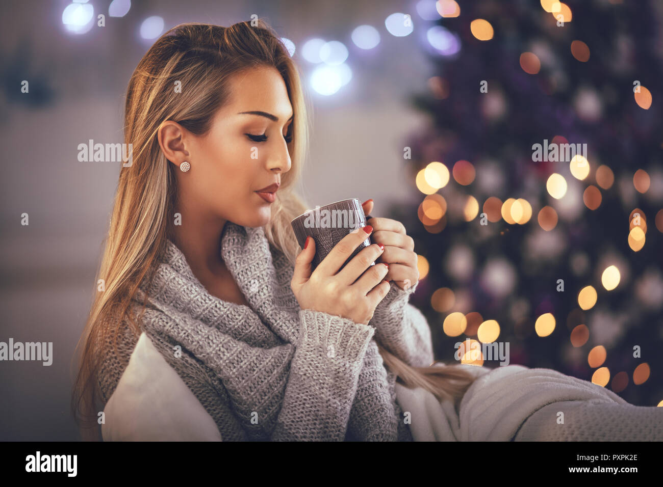 Nette junge Frau mit Tasse Kaffee und Sitzen im Bett mit Weihnachtsbeleuchtung. Stockfoto