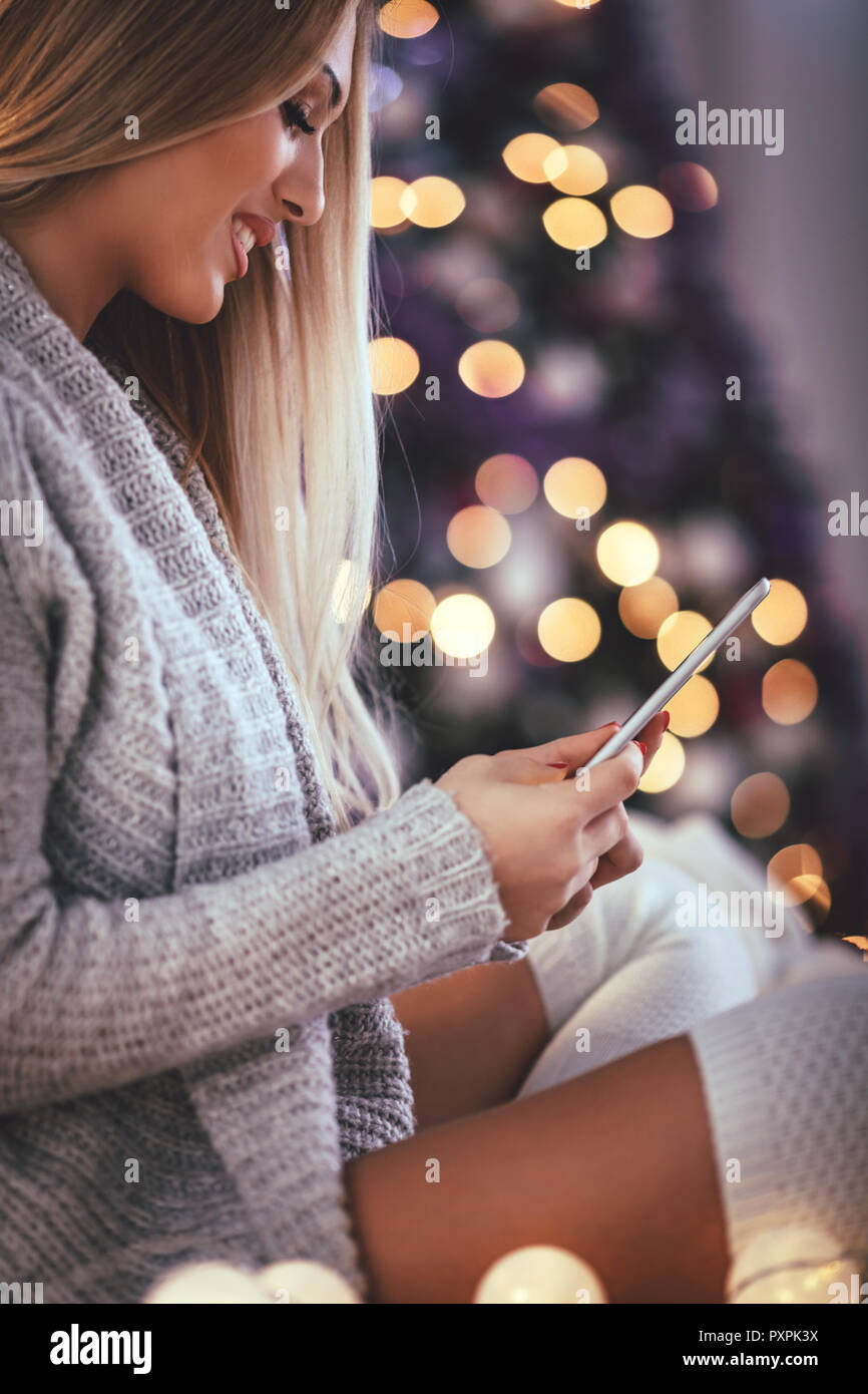 Nette junge lächelnde Frau mit Smartphone und während gemütliche Weihnachten Urlaub zu Hause ein Lächeln auf den Lippen. Stockfoto