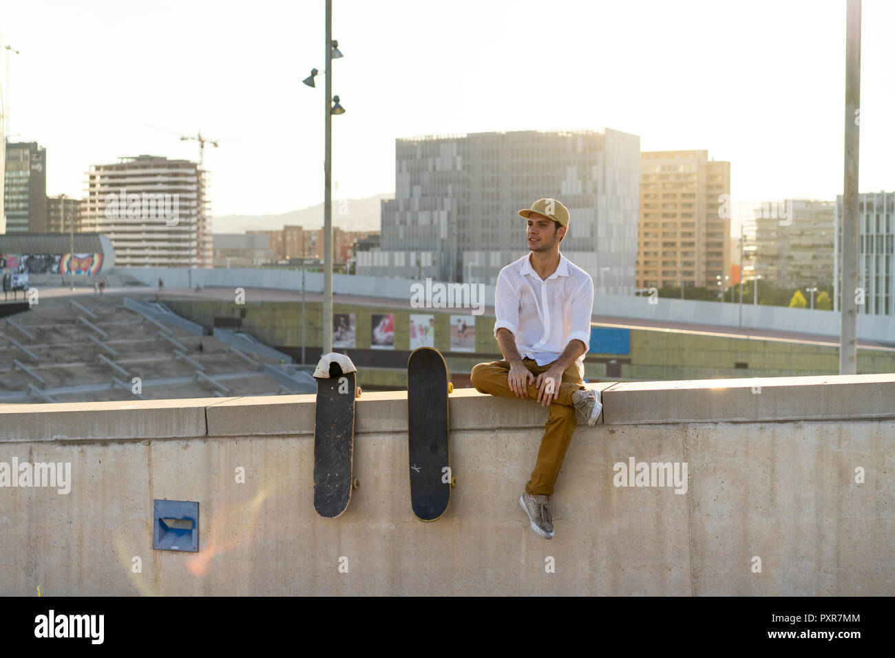 Junge Mann sitzt auf städtischen Wand neben Skateboards bei Sonnenuntergang Stockfoto