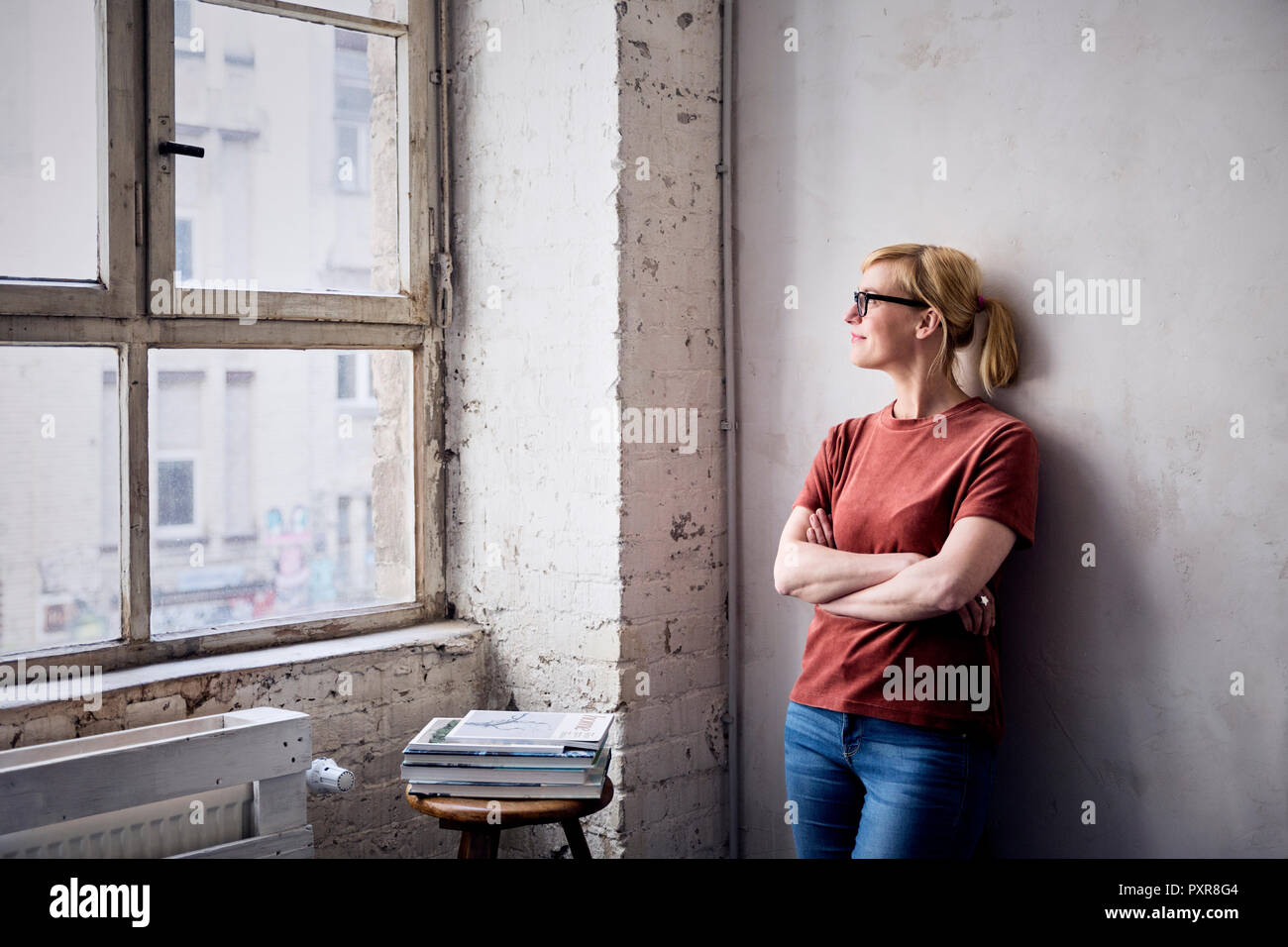 Lächelnde Frau lehnte sich gegen die Wand im Loft auf der Suche durch das Fenster Stockfoto