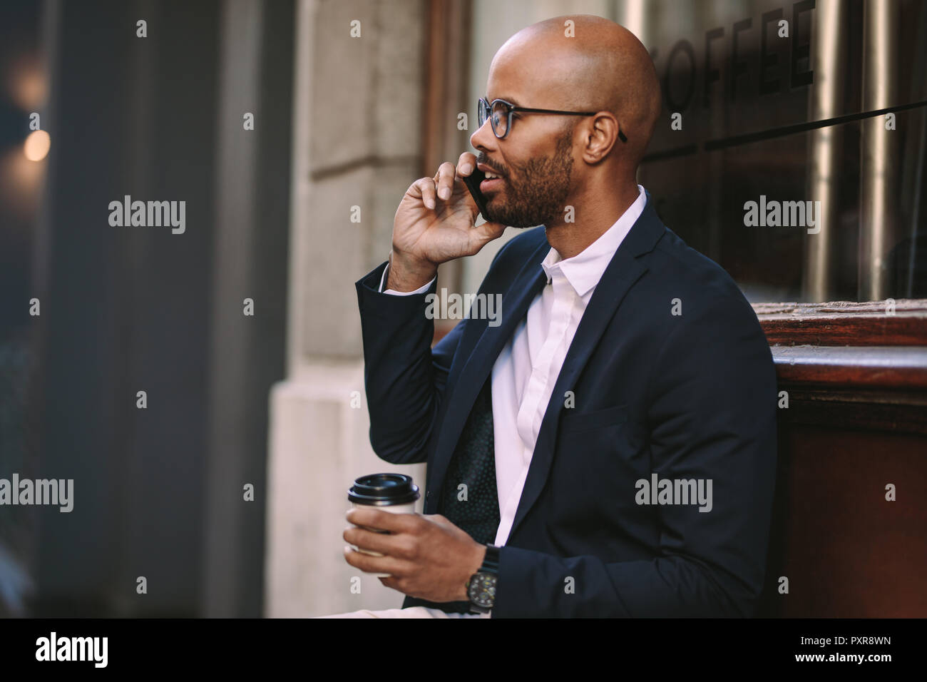 Glücklicher afrikanischer Geschäftsmann, der draußen sitzt und mit einer Tasse Kaffee telefoniert. Geschäftsmann, der sich draußen entspannt und telefoniert. Stockfoto