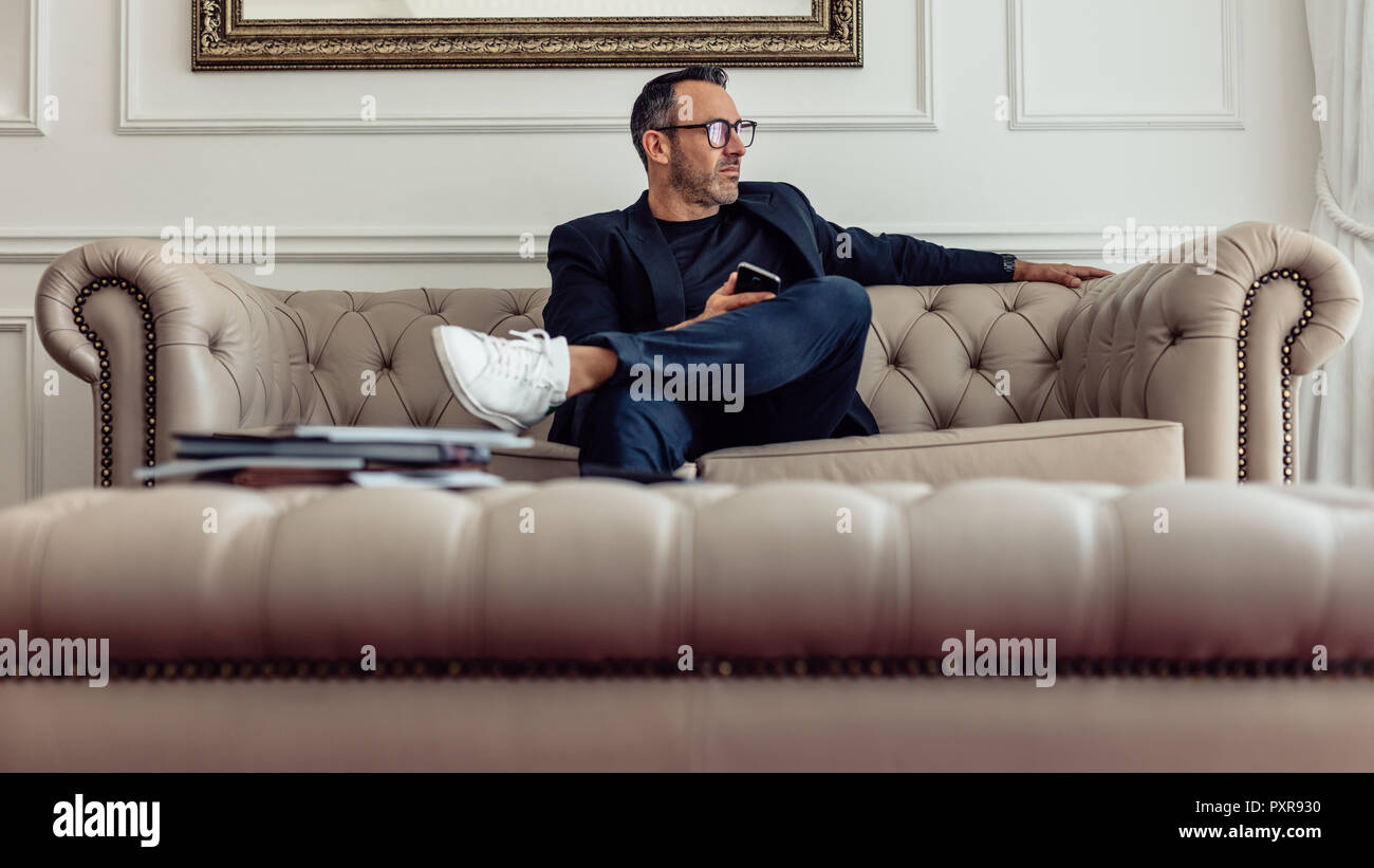 Stilvolle reife Geschäftsmann sitzen auf der Couch im Zimmer und Wegsehen. CEO einer Firma auf Geschäftsreise in einem Hotel zu übernachten. Stockfoto